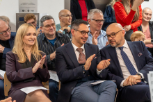 Grupa osób, w tym kobiety i mężczyźni, siedzi w sali podczas formalnego wydarzenia. Uczestnicy ubrani są elegancko w garnitury i stroje biznesowe, część trzyma dokumenty lub programy spotkania, a niektórzy klaszczą.