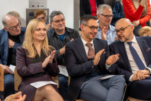 Grupa osób, w tym kobiety i mężczyźni, siedzi w sali podczas formalnego wydarzenia. Uczestnicy ubrani są elegancko w garnitury i stroje biznesowe, część trzyma dokumenty lub programy spotkania, a niektórzy klaszczą.