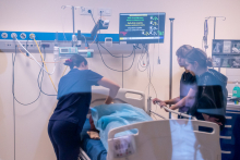 Trzy osoby stoją przy łóżku w sali medycznej. Na łóżku leży fantom przykryty niebieskim materiałem. Jedna osoba pochyla się nad fantomem i wykonuje czynności przy jego klatce piersiowej. Pozostałe osoby obserwują z bliska. Nad łóżkiem wisi monitor z kolorowym tekstem i parametrami. W tle widoczny jest sprzęt medyczny. 