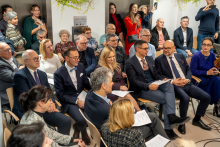 Koncert odbywający się w holu szpitalnym. Publiczność siedzi na krzesłach ustawionych w kilku rzędach, a część osób stoi z tyłu. W pierwszym rzędzie znajdują się osoby w eleganckich strojach, trzymające w dłoniach kartki.