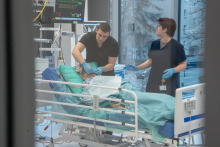 Dwie osoby stoją przy łóżku w sali medycznej. Na łóżku leży fantom przykryty zielonym materiałem. Jedna osoba pochyla się nad fantomem i wykonuje czynności przy głowie, druga stoi obok i trzyma rękę na barierce. Obie osoby mają na dłoniach niebieskie rękawiczki. W tle widać monitor z parametrami i sprzęt medyczny.