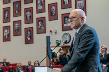 Elegancko ubrany mężczyzna stoi za mównicą i przemawia. W oddali widać słuchające go osoby. Na ścianie auli widać czarno-białe portrety.