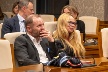 Osoby siedzą w sali konferencyjnej na jasnych, tapicerowanych krzesłach ustawionych w rzędach. Na stole przed nimi leżą dokumenty