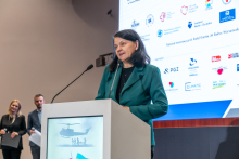 An elegantly dressed woman stands behind a lectern and gives a speech. In the background, there is a multimedia screen displaying various logos.