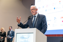 An elegantly dressed man stands behind a lectern and speaks. His right hand is raised. In the background, there is a multimedia screen with various logos.