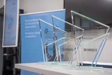 Na białym stole stoją cztery szklane statuetki w kształcie trójkątów. Na każdej widnieje napis informujący o Międzynarodowej Olimpiadzie Wiedzy oraz nazwie uczelni. Statuetki oznaczają miejsca: pierwsze, drugie i dwa trzecie. W tle widać niebieski baner z nazwą Warszawskiego Uniwersytetu Medycznego oraz fragment ekranu prezentacyjnego.