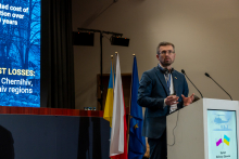 An elegantly dressed man stands behind a lectern and gives a speech. In the background, you can see part of a multimedia screen and three flags.