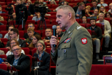 A man dressed in uniform with a microphone in his hand. In the background, other people are sitting on red chairs.