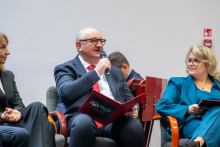 Three people are sitting on chairs in a conference room during a panel discussion. One person is holding a microphone and a red folder with the words “SAVE A LIFE” written on it. Everyone is dressed elegantly.