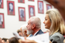 Several people are sitting in an auditorium during a conference. In the foreground, you can see people in elegant attire, including a gray suit. In the background, rows of red frames with photos and inscriptions hang on the wall.