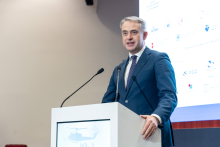 An elegantly dressed man stands behind a lectern and gives a speech. A multimedia screen can be seen in the background.