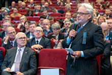 Elegancko ubrany mężczyzna z mikrofonem w ręce, za nim - wiele osób siedzących na czerwonych krzesłach.