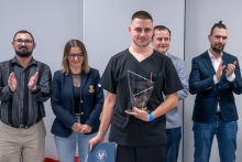 Pięć osób stoi w rzędzie w sali. Osoba na środku, Filip Walkowiak, trzyma otwarty dyplom, szklaną statuetkę i torbę z logo uczelni. Pozostałe osoby stoją obok w eleganckich strojach. 