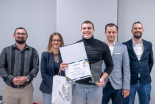 Pięć osób stoi w rzędzie w sali. Osoba na środku, Piotr Adamczyk, trzyma otwarty dyplom, szklaną statuetkę i torbę z logo uczelni. Pozostałe osoby stoją obok w eleganckich strojach. 