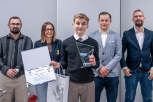 Pięć osób stoi w rzędzie w sali. Osoba na środku, Tomasz Czarnowski, trzyma otwarty dyplom, szklaną statuetkę i torbę z logo uczelni, co wskazuje na odbiór nagrody. Pozostałe osoby stoją obok w eleganckich strojach. 