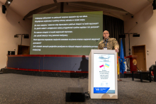A man in uniform stands behind a lectern and speaks. In the background, there is a multimedia screen and three flags.