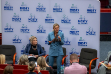 Two people are participating in a panel discussion against a backdrop with the words “Bridges to Health.” One person is seated and holding notes, while the other—the Minister of Education—is standing with a microphone and tablet. In the foreground, several people can be seen in the auditorium, some of them taking photos. 