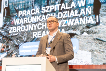 An elegantly dressed man stands behind a lectern and gives a speech. In the background, there is a multimedia screen displaying a presentation.