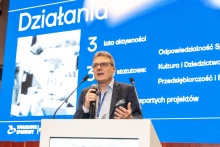 An elegantly dressed man stands behind a lectern and gives a speech. In the background, there is a multimedia screen displaying a presentation.