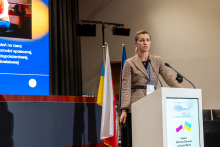An elegantly dressed woman stands behind a lectern and gives a speech. In the background, you can see part of a multimedia screen and three flags.