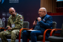 Two men in an auditorium during a panel discussion. One is wearing a military uniform with a patch and is holding a microphone. The other, an elegantly dressed man, Prof. Robert Gałązkowski, is also holding a microphone. 