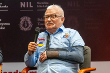 President Lech Wałęsa during a panel discussion. He is holding a microphone in his right hand and speaking. 