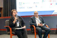 Two people in discussion. On the left sits a woman with a microphone in her hand; on the right, an elegantly dressed man sits and listens.