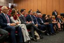 Ten people are sitting in the front row of the auditorium during a conference. They are dressed elegantly. In the background, you can see more rows of participants.