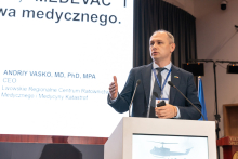 An elegantly dressed man giving a speech. He is standing behind a lectern. His right hand is raised.