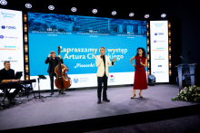 Cztery osoby na scenie podczas występu – jedna gra na keyboardzie, druga na kontrabasie, a dwie stoją w centrum, z czego jedna śpiewa lub przemawia do mikrofonu. W tle duży ekran.