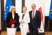 Three people are standing in front of the flags of Poland and the European Union in an elegant room, one of them holding a burgundy briefcase and a bag. On the right-hand side, there is a lectern with the inscription “Ministry of Health.”