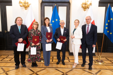 Six people are standing in an elegant room in front of the flags of Poland and the European Union, four people are holding burgundy briefcases and white bags. On the right-hand side, there is a lectern with the inscription “Ministry of Health.”