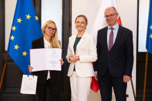 Three people are standing in front of the flags of Poland and the European Union in an elegant room, one of them holding an open document and a bag. On the right-hand side, there is a lectern with the inscription “Ministry of Health.”
