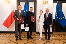Four people are standing in an elegant room in front of the flags of Poland and the European Union, two people are holding burgundy briefcases and white bags. On the right-hand side, there is a lectern with the inscription “Ministry of Health.”