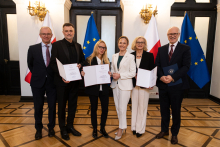 Six people are standing in an elegant room in front of the flags of Poland and the European Union, four people are holding open briefcases. On the right-hand side.