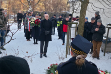 Grupa osób stoi na ośnieżonym terenie podczas uroczystości upamiętniającej. Na środku stoi mężczyzna w ciemnym płaszczu z rękami splecionymi przed sobą. Za nim kobieta trzyma duży bukiet biało‑czerwonych kwiatów, a obok stoją funkcjonariusze i uczestnicy wydarzenia. Po prawej stronie kilka osób obserwuje przebieg uroczystości. 