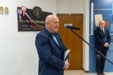 Mężczyzna w garniturze stoi przy mikrofonie, trzymając dokumenty w dłoni; w tle widoczna jest tablica pamiątkowa z medalionem.