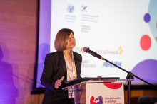 Elegancko ubrana kobieta stoi przy mównicy i mówi do mikrofonu. Patrzy przed siebie. W tle widać duży ekran z logotypami instytucji.