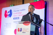 Elegancko ubrany mężczyzna stoi przy mównicy i mówi do mikrofonu. W tle widać duży baner konferencyjny z kolorową grafiką i tekstem.