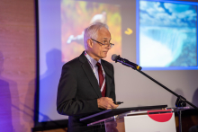 Starszy, elegancko ubrany mężczyzna stoi przy mównicy i mówi do mikrofonu. Trzyma pilota do zmiany slajdów w jednej ręce. Patrzy w bok i ma spokojny wyraz twarzy. Za nim wyświetlam slajdy z kolorowymi zdjęciami.