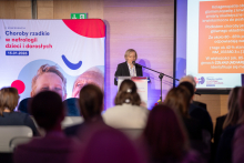 Elegancko ubrana kobieta stoi przy mównicy i mówi do mikrofonu. Patrzy w stronę publiczności. Przed nią siedzi kilkanaście osób, które słuchają wystąpienia. Obok niej stoją baner konferencji oraz ekran z widocznym slajdem z tekstem.