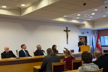 Spotkanie w sali obrad. Sześć osób siedzi za długim stołem, a jedna osoba stoi przy mównicy i przemawia do zebranych. Wszyscy patrzą w stronę występującego i zachowują spokojną postawę. Obok stołu stoją flagi. 