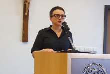 Elegancko ubrana kobieta stoi za mównicą i przemawia.