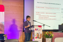 Kobieta stojąca za mównicą, która przemawia. W tle widać ekran z wyświetlanym slajdem.