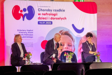 Trzy osoby siedzące na krzesłach. Za nimi widać duży baner konferencyjny z kolorową grafiką i tekstem.