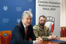 Dwóch mężczyzn siedzi przy stole w sali konferencyjnej. Jeden nosi garnitur z białą koszulą i krawatem, drugi ma mundur w barwach wojskowych. Na stole leży granatowa teczka. W tle widać niebieską tablicę z napisem „Warszawski Uniwersytet Medyczny” oraz biały baner z logo i napisem „Centralny Pododdział Kontrterrorystyczny Policji BOA”.