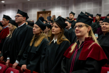 Kilkudziesięciu doktorów i doktorów habilitowanych w togach stoi w rzędach podczas uroczystości. Osoby patrzą przed siebie.