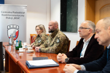 Cztery osoby siedzą przy stole w sali konferencyjnej. Dwie osoby noszą mundury w barwach wojskowych, a dwie osoby mają eleganckie ubrania. W tle znajduje się biały baner z logo i napisem „Centralny Pododdział Kontrterrorystyczny Policji BOA”. 