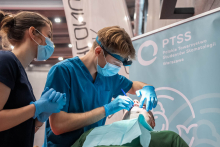 Trzy osoby biorą udział w pokazie stomatologicznym. Jedna osoba leży na fotelu, a dwie osoby w rękawiczkach pochylają się nad jej ustami i wykonują zabieg. Jedna z nich używa narzędzia stomatologicznego, a druga asystuje z boku. 