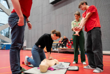 Czworo młodych osób uczestniczy w ćwiczeniach pierwszej pomocy na sali sportowej. Jedna osoba klęczy przy fantomie i wykonuje uciśnięcia klatki piersiowej. Pozostałe osoby stoją obok i udzielają wskazówek. W tle kilka osób siedzi na ławce i obserwuje zajęcia. 